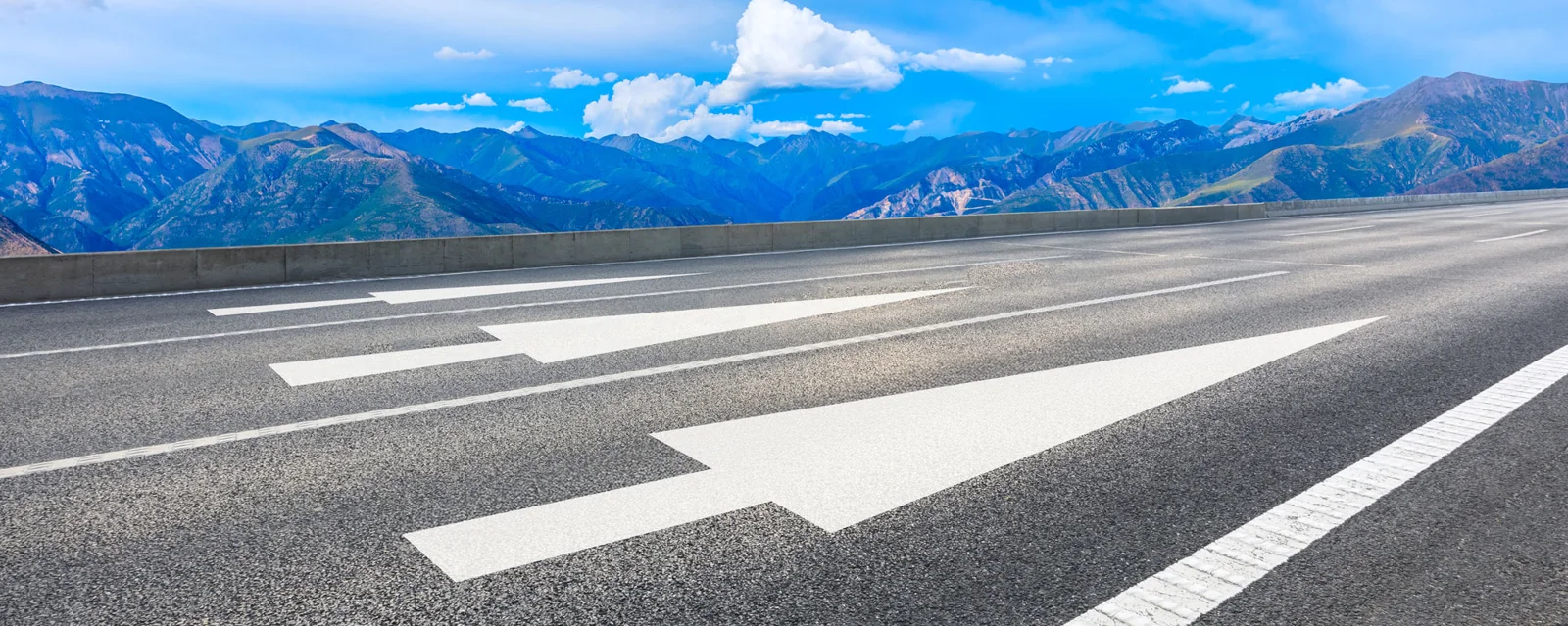 Asphaltstraße mit weißen Richtungspfeilen vor einer Berglandschaft